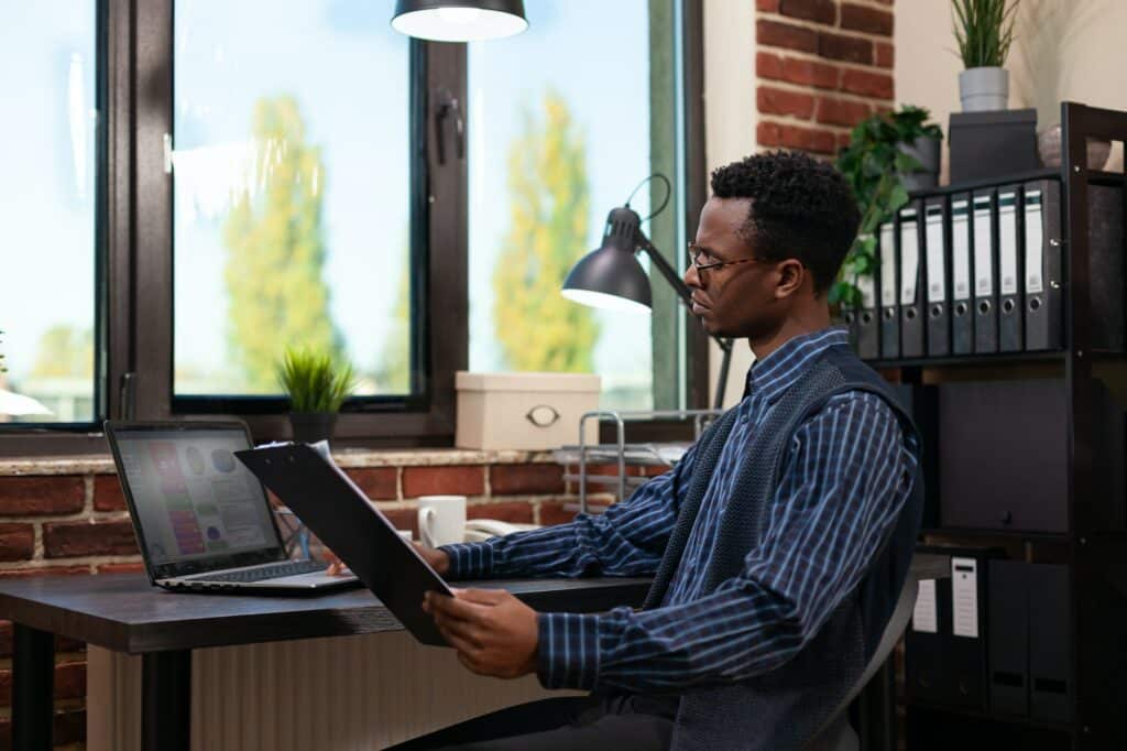 Entrepreneur looking at laptop screen with sales results holding clipboard with bussiness charts