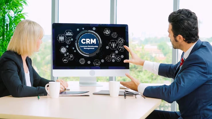 CRM consultant explaining customer relationship management dashboard features to client on large monitor