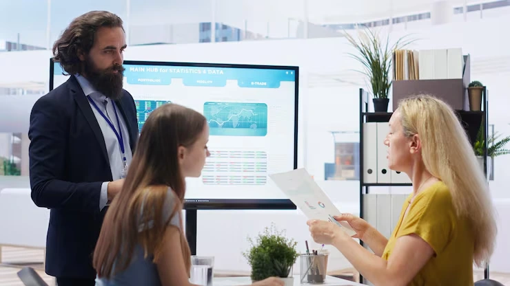Analytics consultant presenting campaign performance data and metrics on large monitor to two business clients