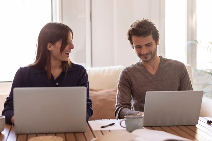 Two marketers working on laptops crafting email campaigns at wooden table in bright office