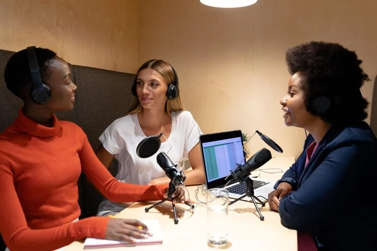 Podcast host interviewing two business clients in studio with recording equipment and audio editing software visible